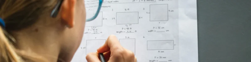 greg-rosenke-DJWvzY4pkGU-unsplash-1 Uma criança com cabelos presos e óculos está concentrada em resolver um exercício de matemática em uma folha de papel. A imagem mostra a visão de trás, com a criança segurando um lápis e escrevendo as respostas. O exercício envolve cálculo de perímetro e medidas desconhecidas de retângulos. O ambiente tem uma iluminação suave, e a mesa cinza contrasta com o papel branco da atividade.